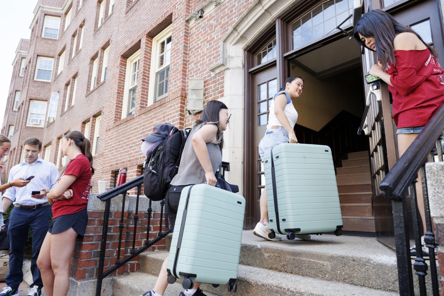 Two students carry suitcases up the stairs to an open dorm door.