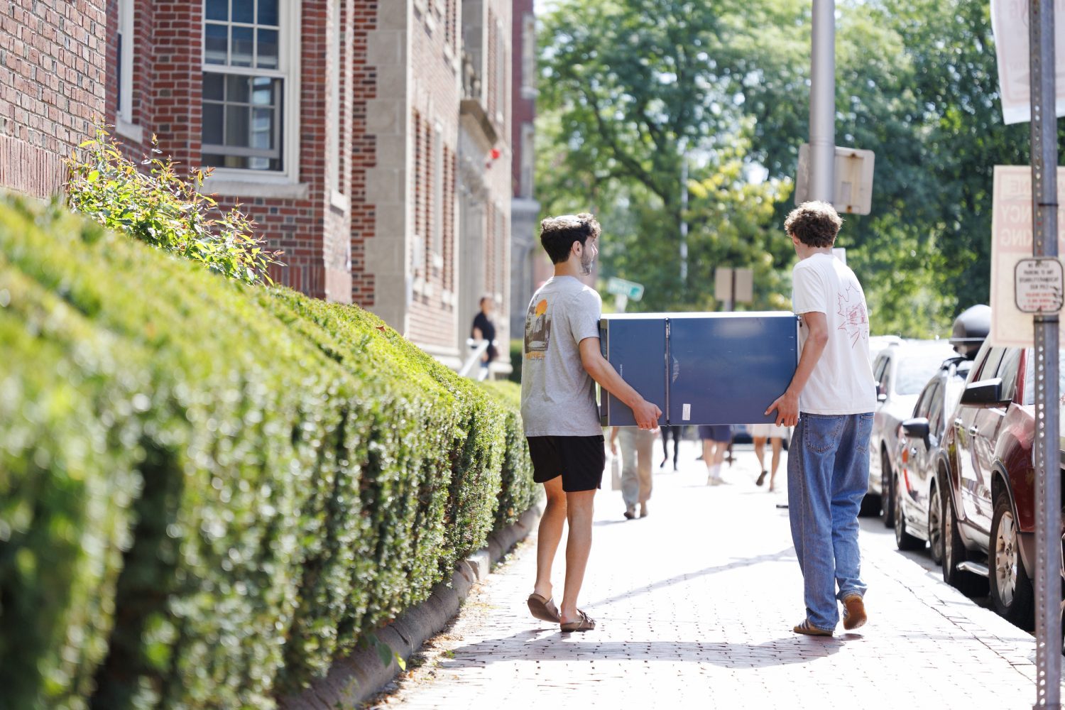 Two people carry a mini fridge down the sidewalk.