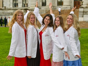 Five women wearing their new white coats pose for a photo with their arms outstretched.
